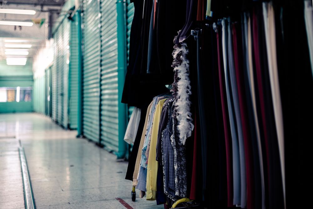 Clothing hangs on racks in a narrow, brightly lit hallway with teal storage units along the walls.