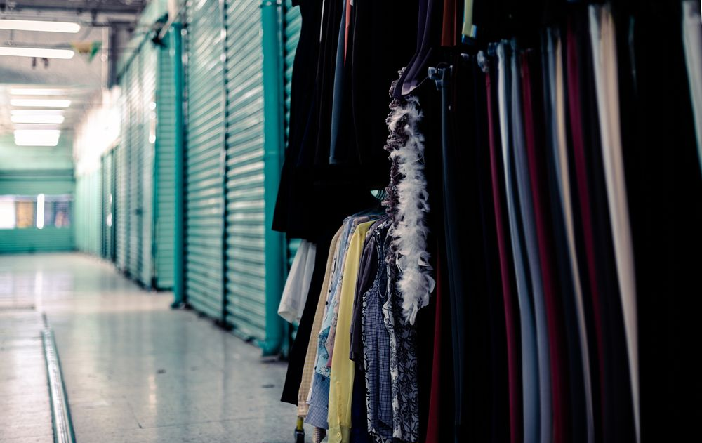 A rack of assorted clothes and a white feather boa hangs in a corridor lined with teal-colored metal shutter doors.