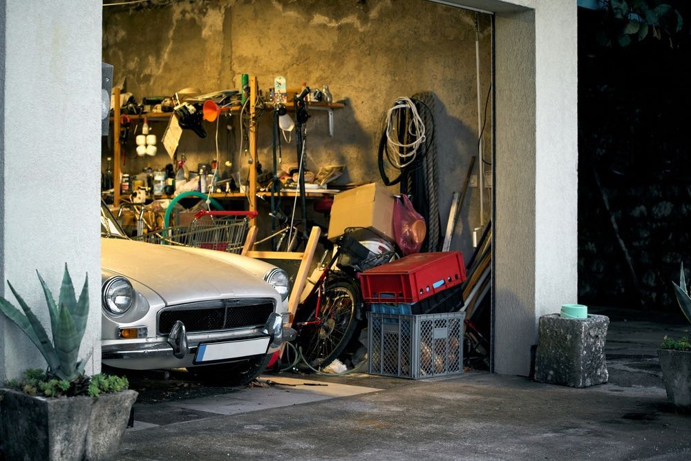 Silver classic car parked in a cluttered garage with tools, crates, and storage items surrounding it