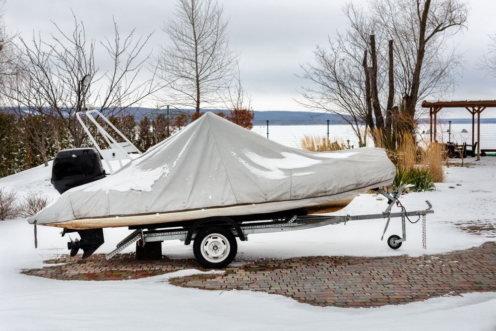 Boat covered with a tarp on a trailer, parked on snow, near a lake.