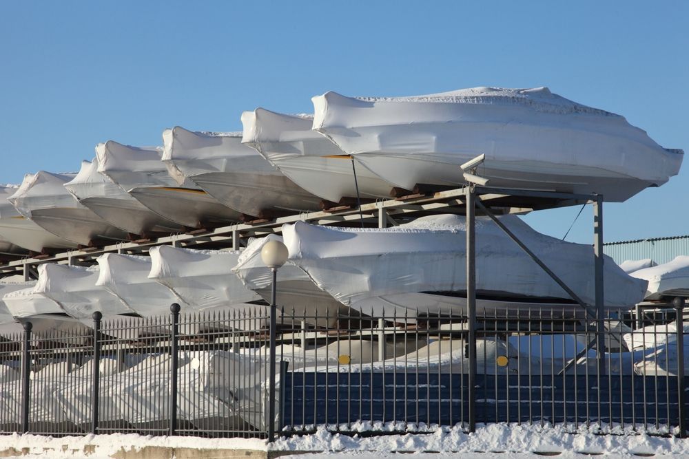 Boats covered in white tarps stacked on racks behind a metal fence, snowy ground, blue sky.