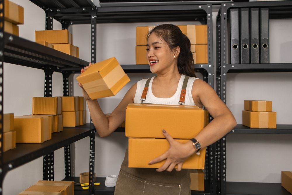 Woman in a storage room holding stacked cardboard boxes on metal shelves