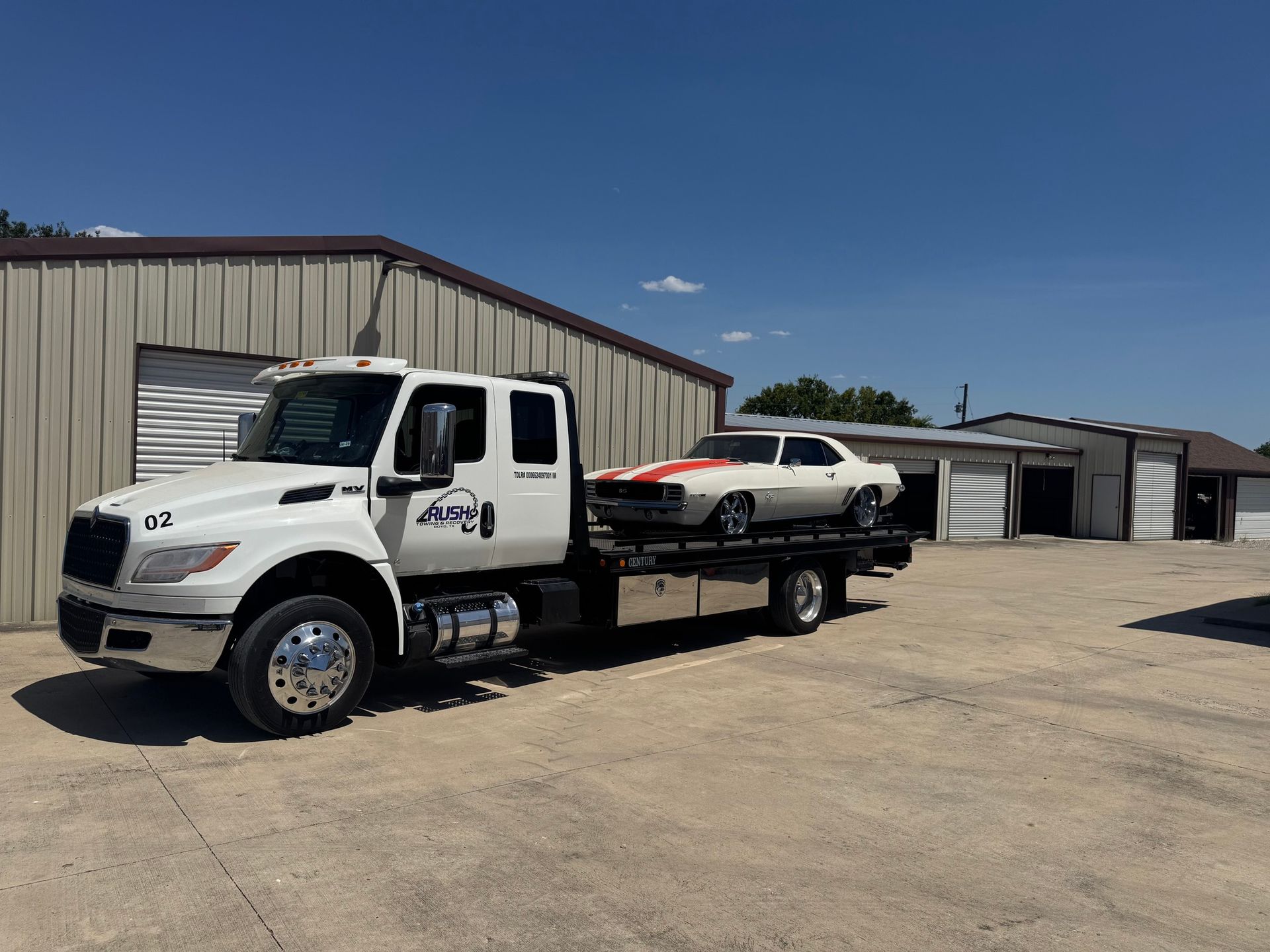White tow truck carrying a white classic car with red stripes; parked outside a brown building on a sunny day.