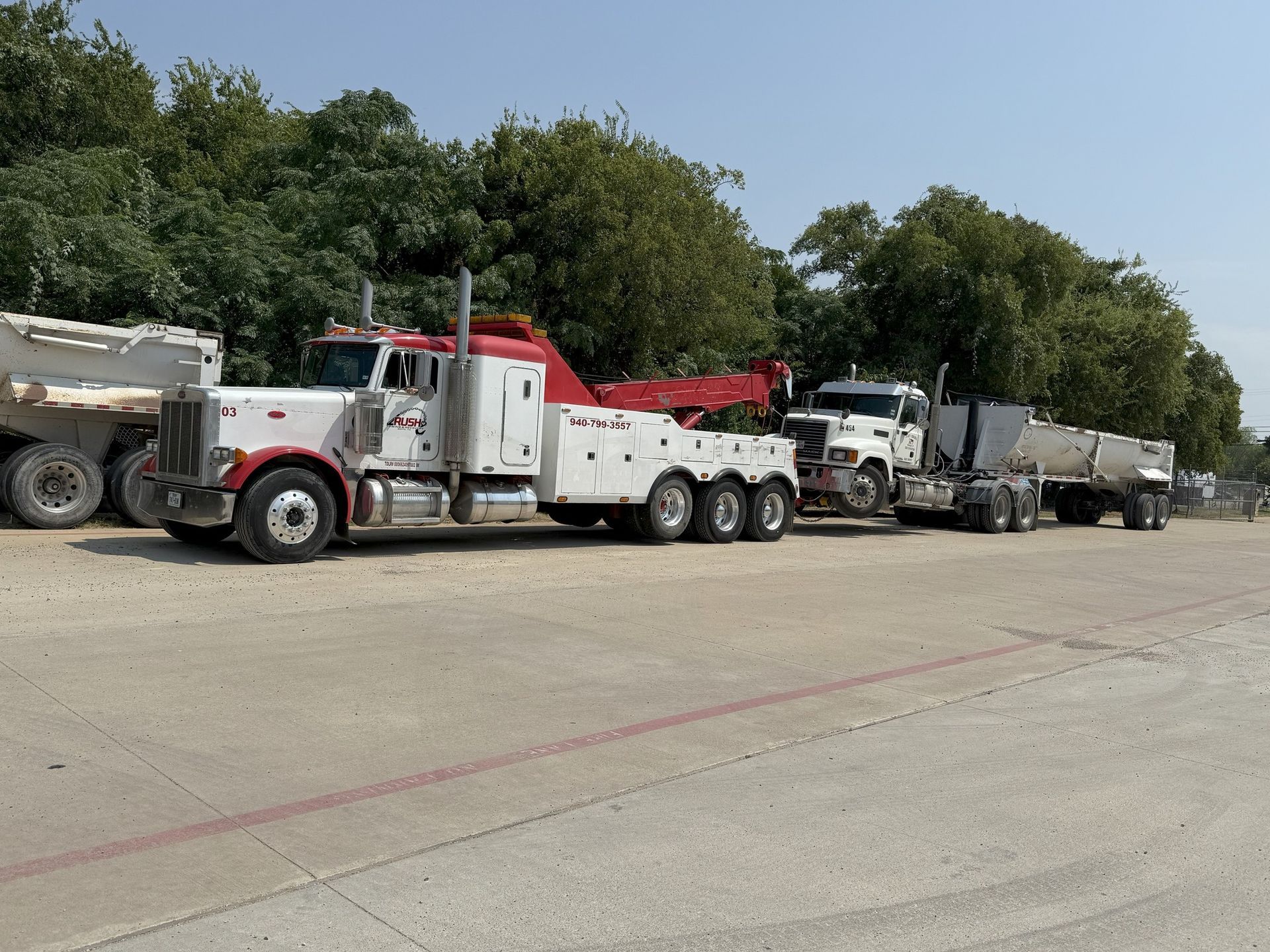 White and red tow truck towing a truck and trailer on a paved lot; trees in the background.