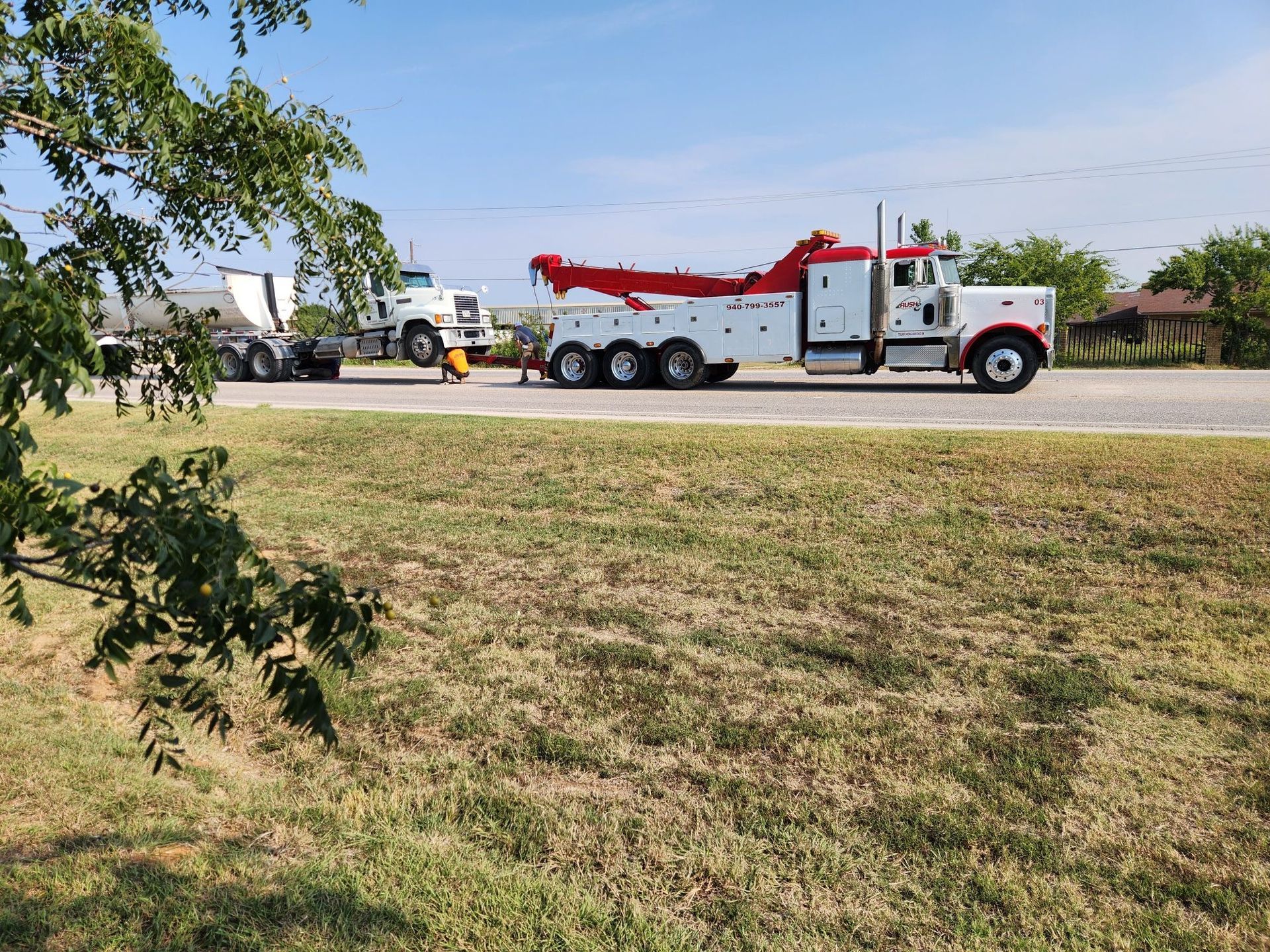 White tow truck towing a white tanker truck on a sunny day. Green trees and brown grass in the foreground.