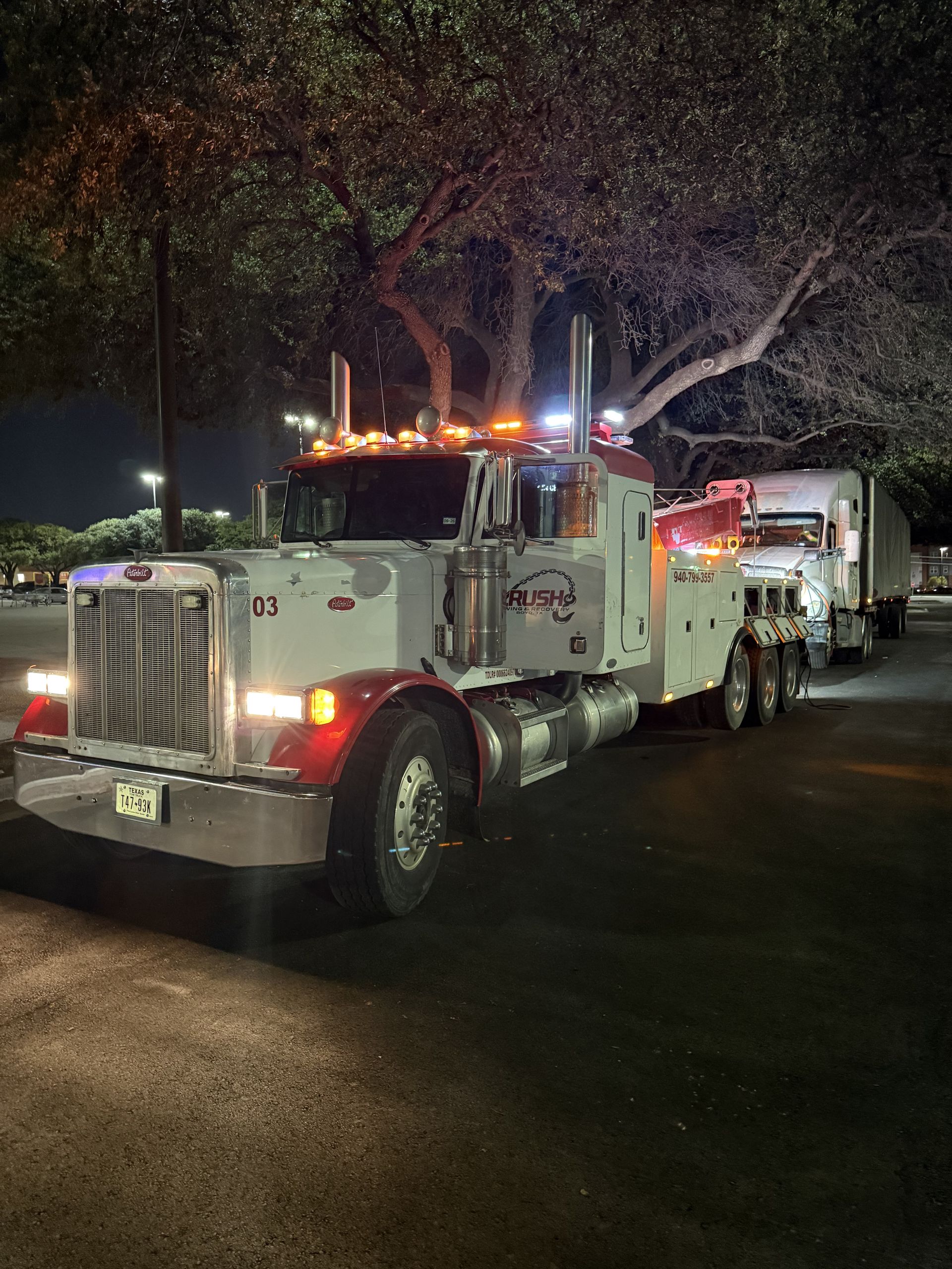 Tow truck with red and white paint parked at night under a tree, lights on.