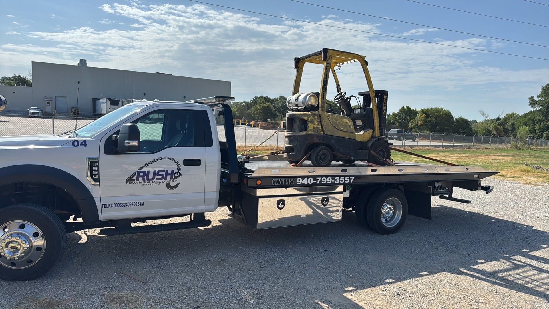 Tow truck transporting a yellow forklift on its flatbed outdoors on a sunny day.