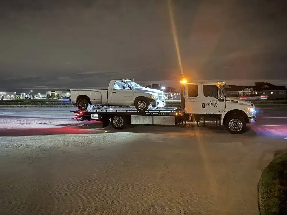 White pickup truck being towed by a tow truck on a road at night; flashing lights.