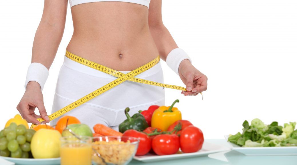 Image of a woman holding a measuring tape around her waist in front of a table full of various nutritious foods