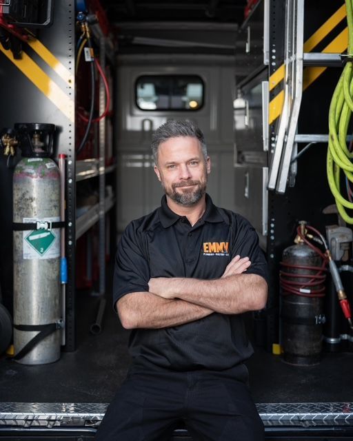 Man standing with arms crossed inside a fire truck, wearing a black uniform with hoses and equipment nearby