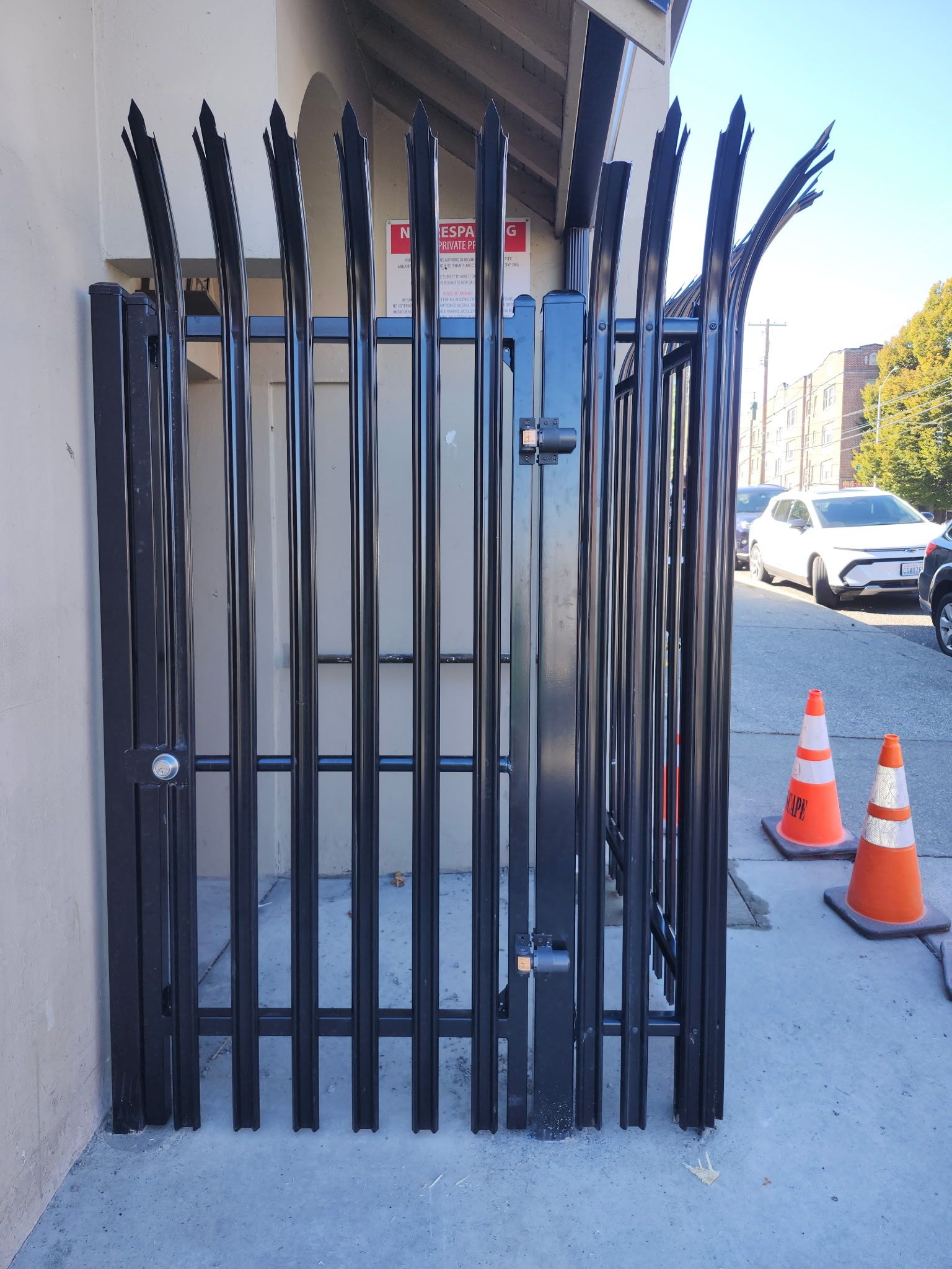 Black metal security gate blocking a walkway beside a building, with traffic cones and parked cars nearby