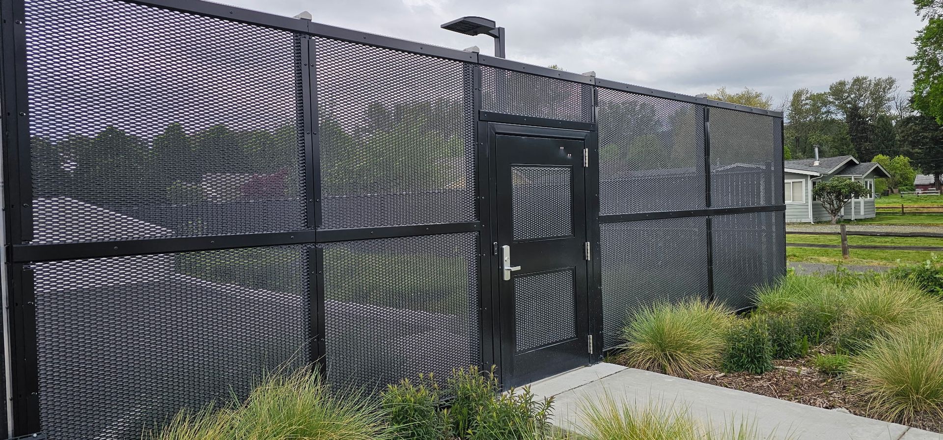 Black chain-link fence with a gate beside a sidewalk and grassy landscaping under cloudy skies