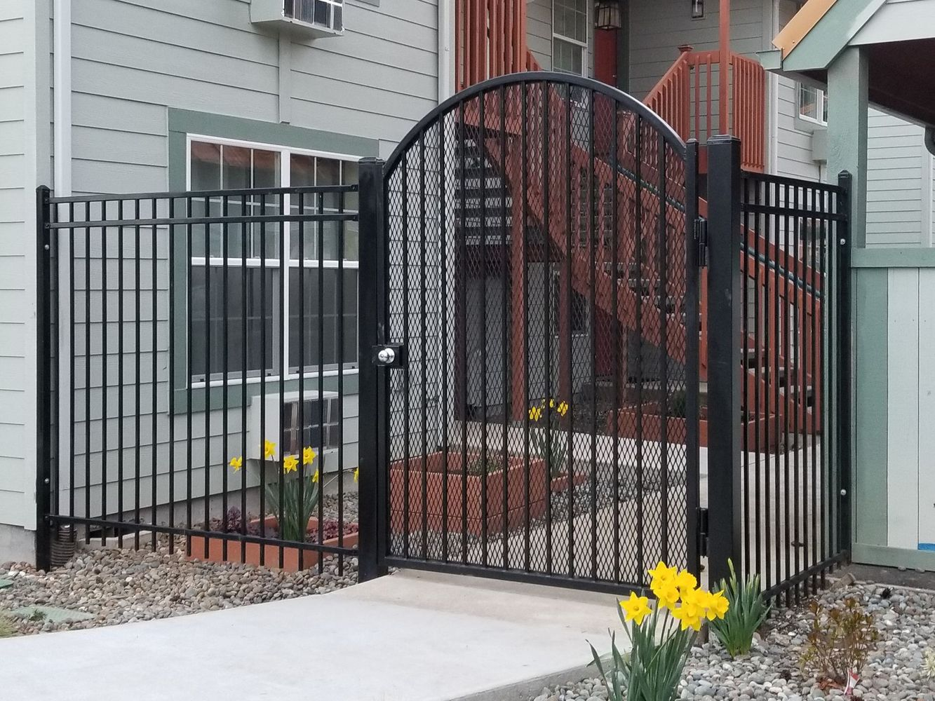 Black metal garden gate and fence with arched top in front of a house, with yellow flowers by the walkway