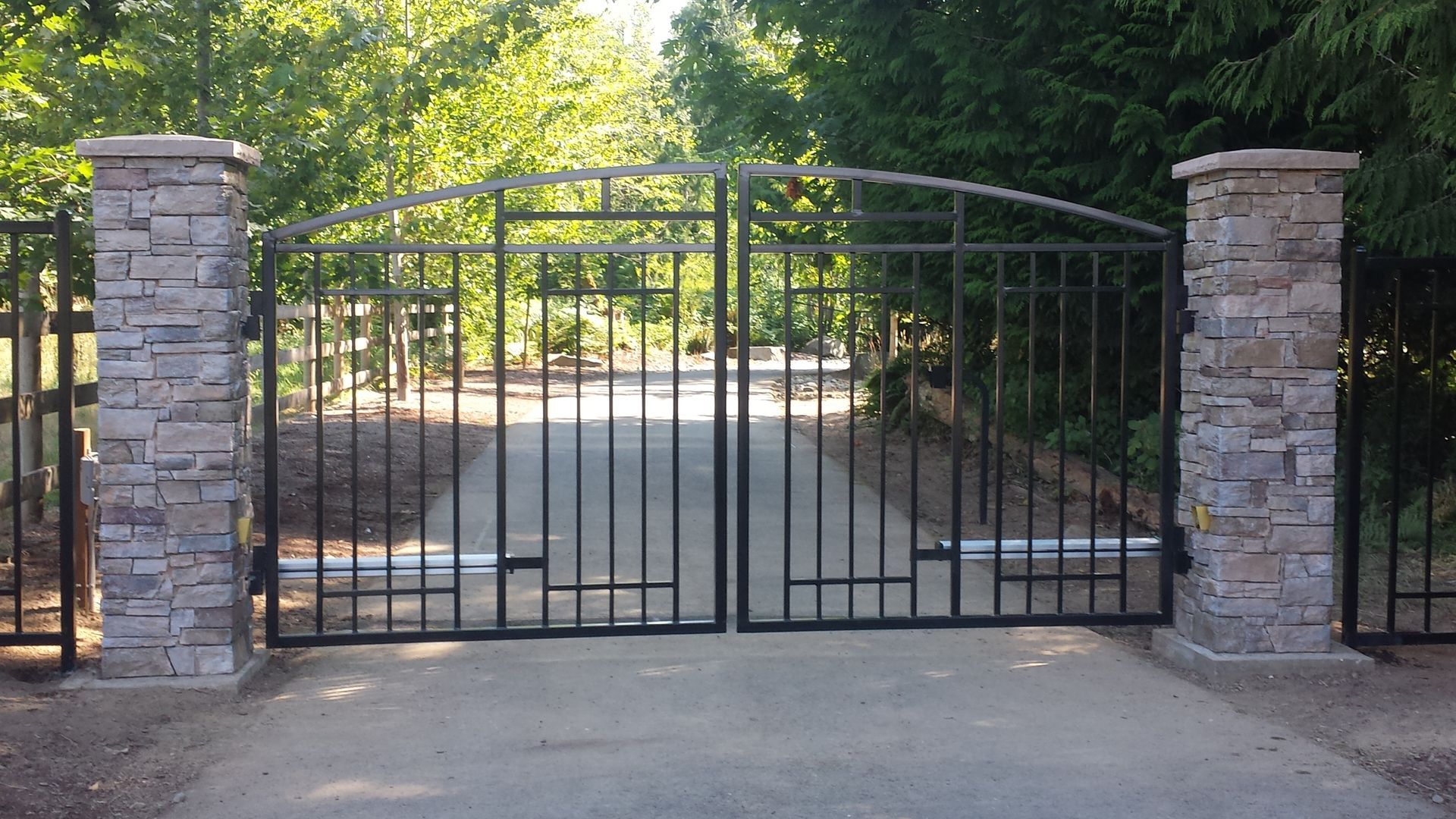 Closed black metal driveway gate between stone pillars, blocking a sunlit driveway