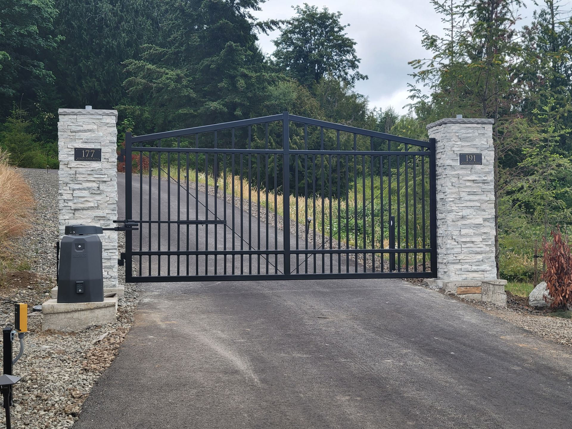 Black metal driveway gate between two stone pillars at the end of a paved drive.