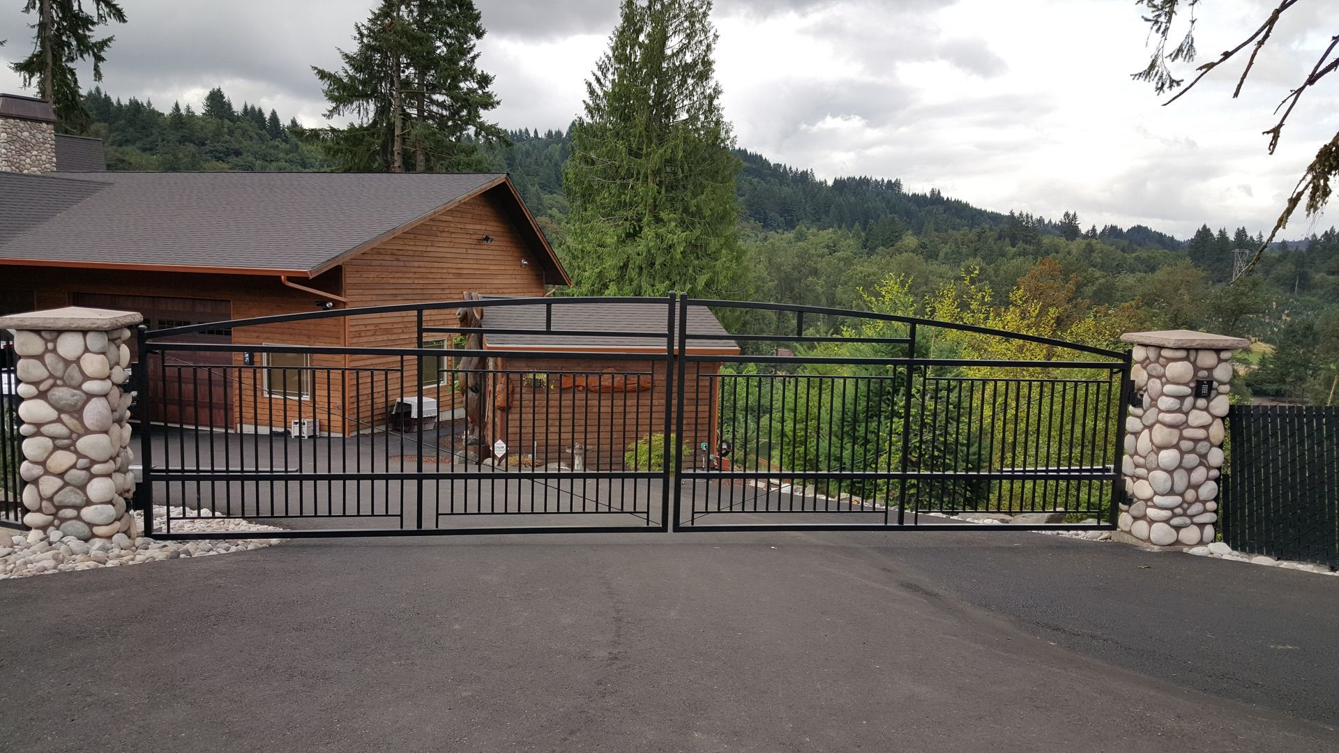 Stone-and-iron driveway gate in front of a wooden house with trees and hills in the background
