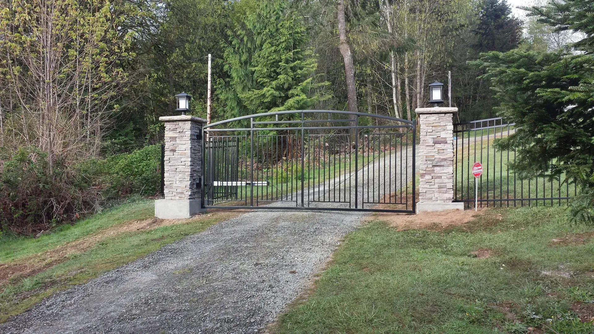 Gravel driveway leading to a closed wrought-iron gate between stone pillars in a wooded area
