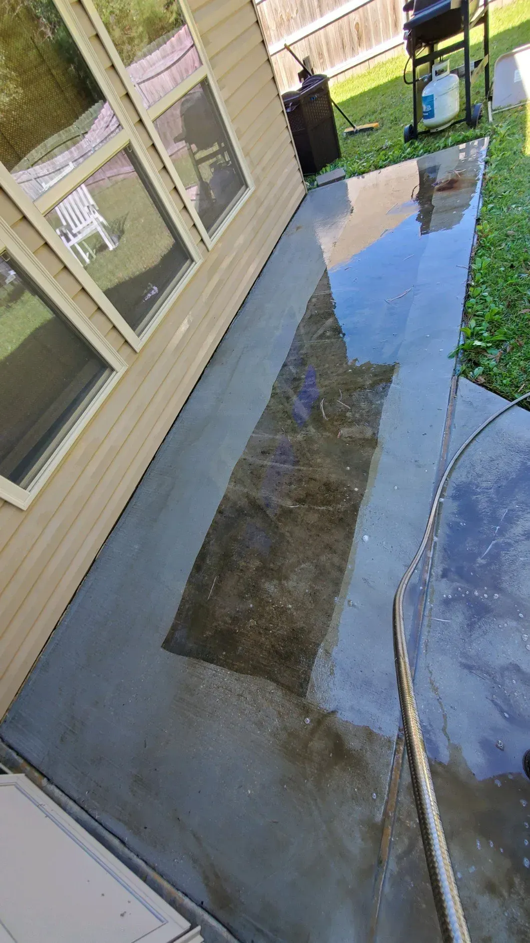 A partial view of a beige house with a concrete patio undergoing pressure washing, showing a clean, wet strip of concrete.