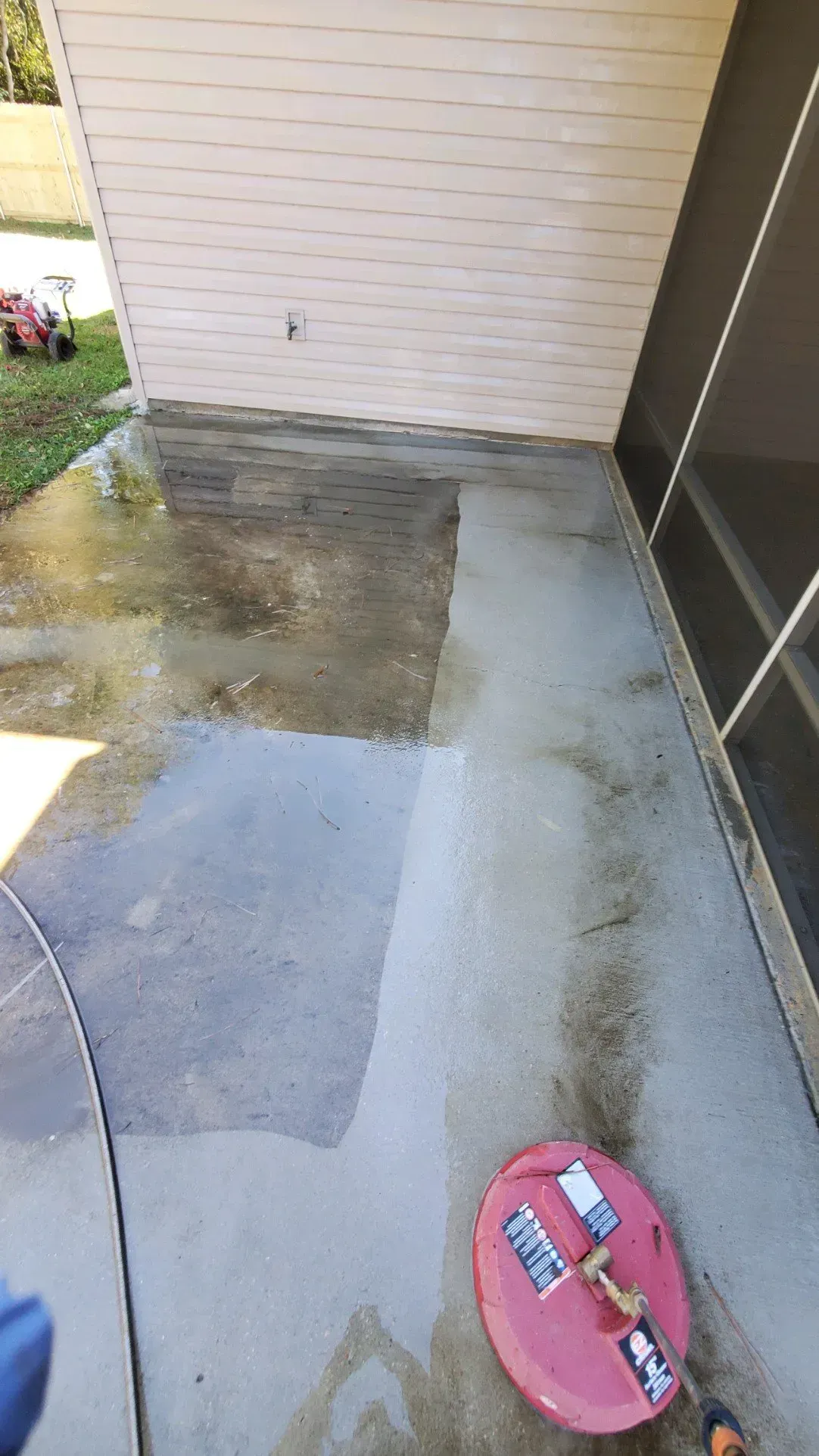 A red circular surface cleaner on a concrete patio, showing a half-cleaned section beside a dirty area.