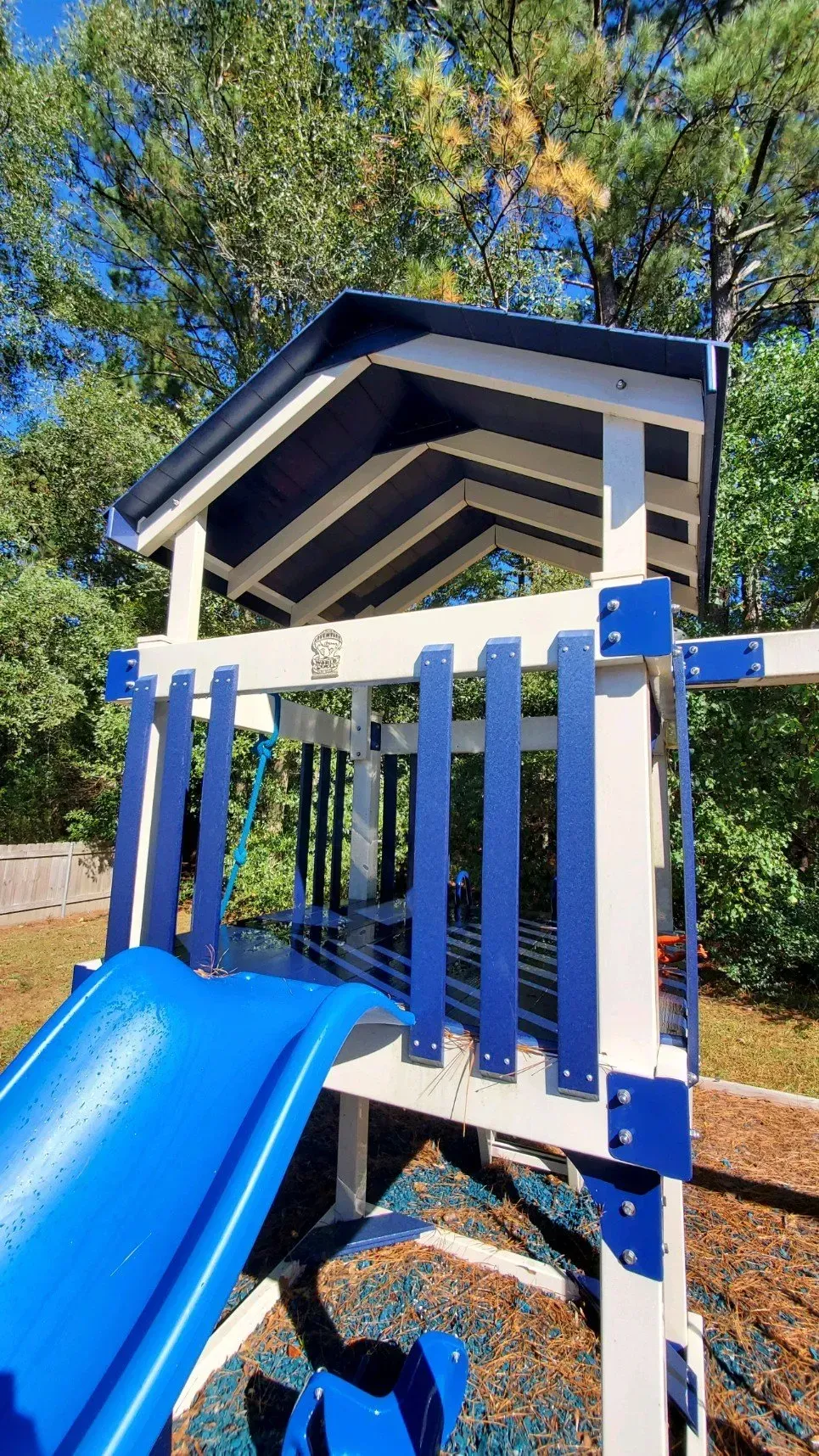 A blue and white backyard playset with a gabled roof and slide, positioned outdoors with green trees in the background.