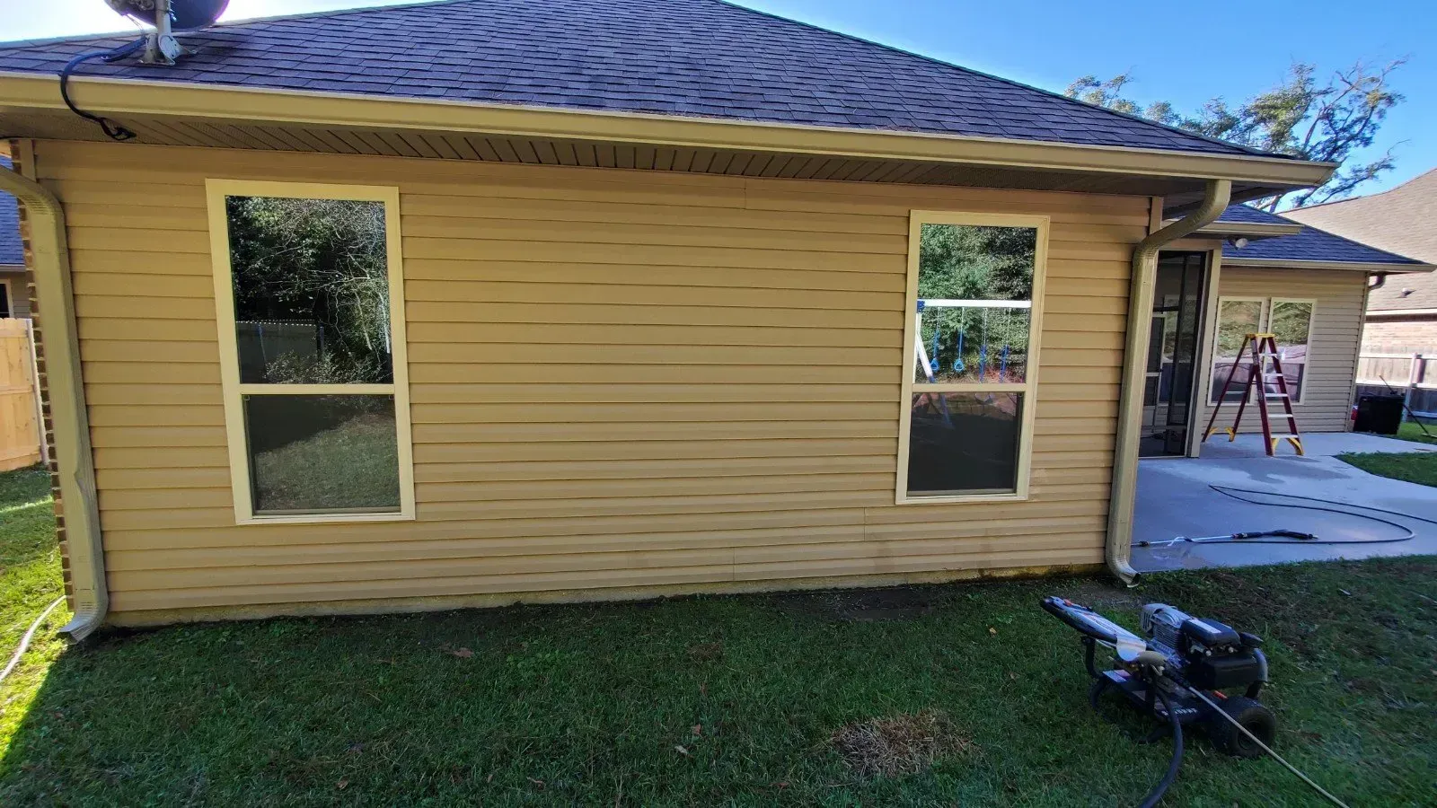 A tan house exterior with two windows, a dark roof, and a pressure washer on the grass in front.