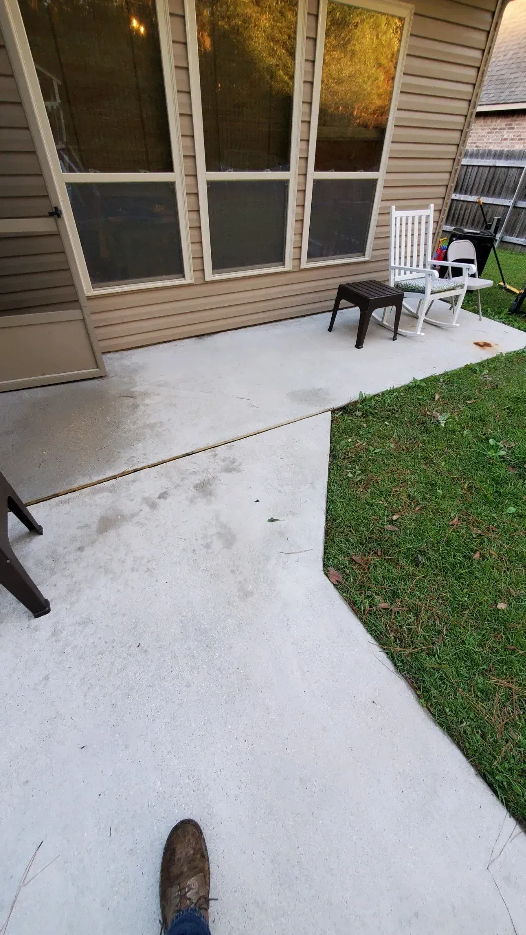 A light-colored concrete patio outside a house with tan siding, featuring a small black stool and white chairs.