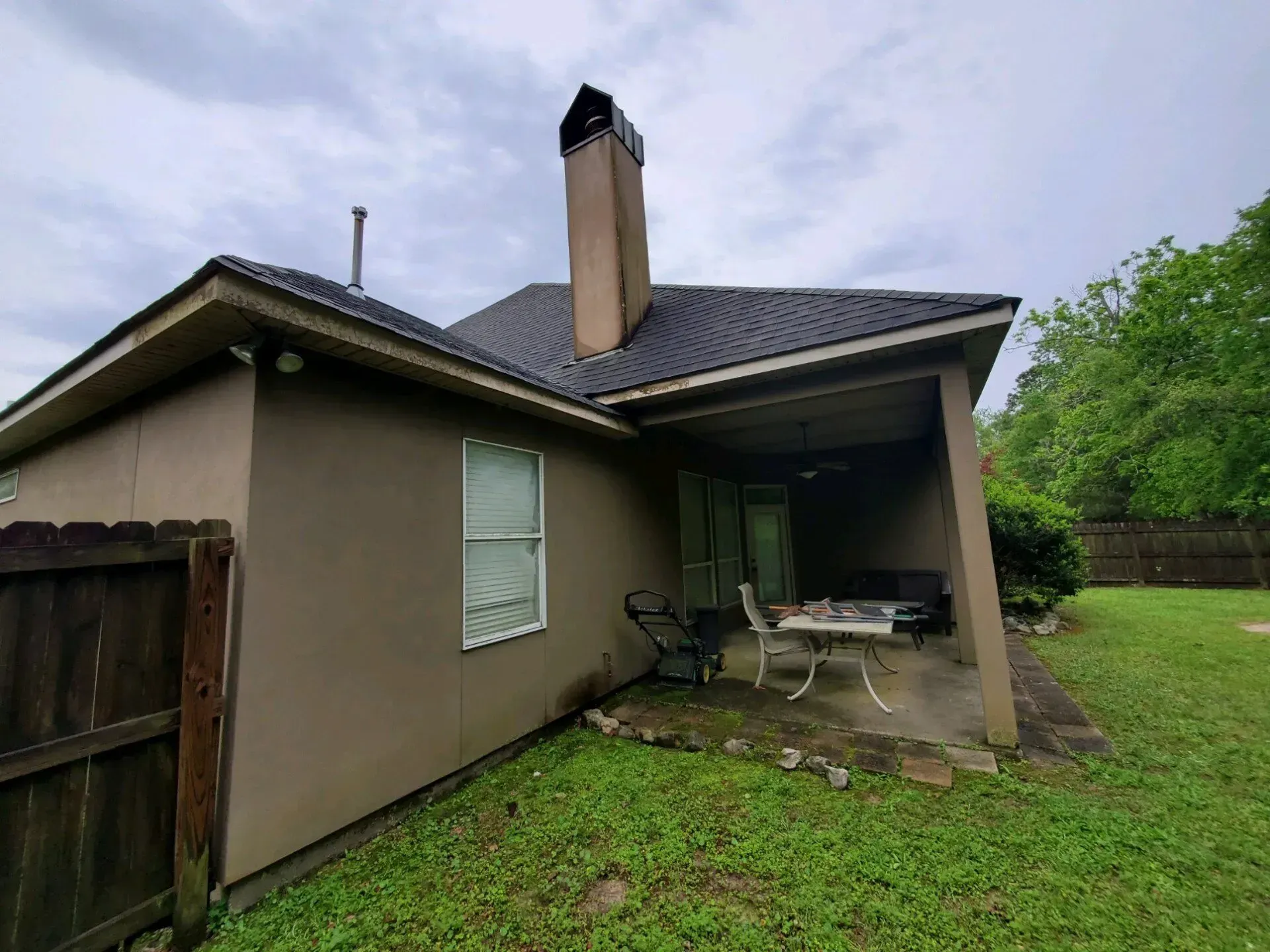A side view of a beige house with a chimney and a covered patio containing a table and chairs, set in a grassy yard.