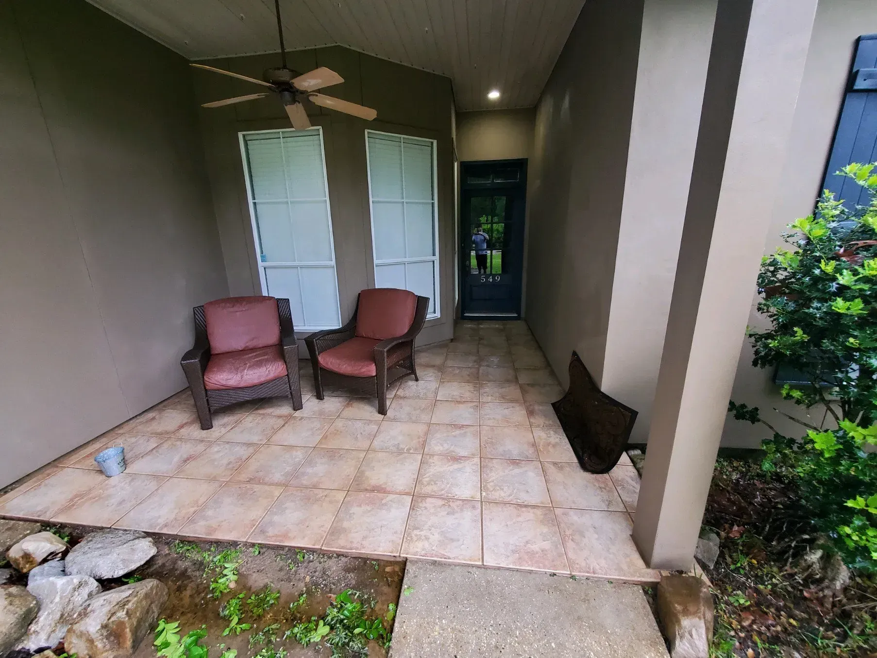 A tiled front porch with two cushioned chairs, a ceiling fan, two windows, and a dark front door under a roof overhang.