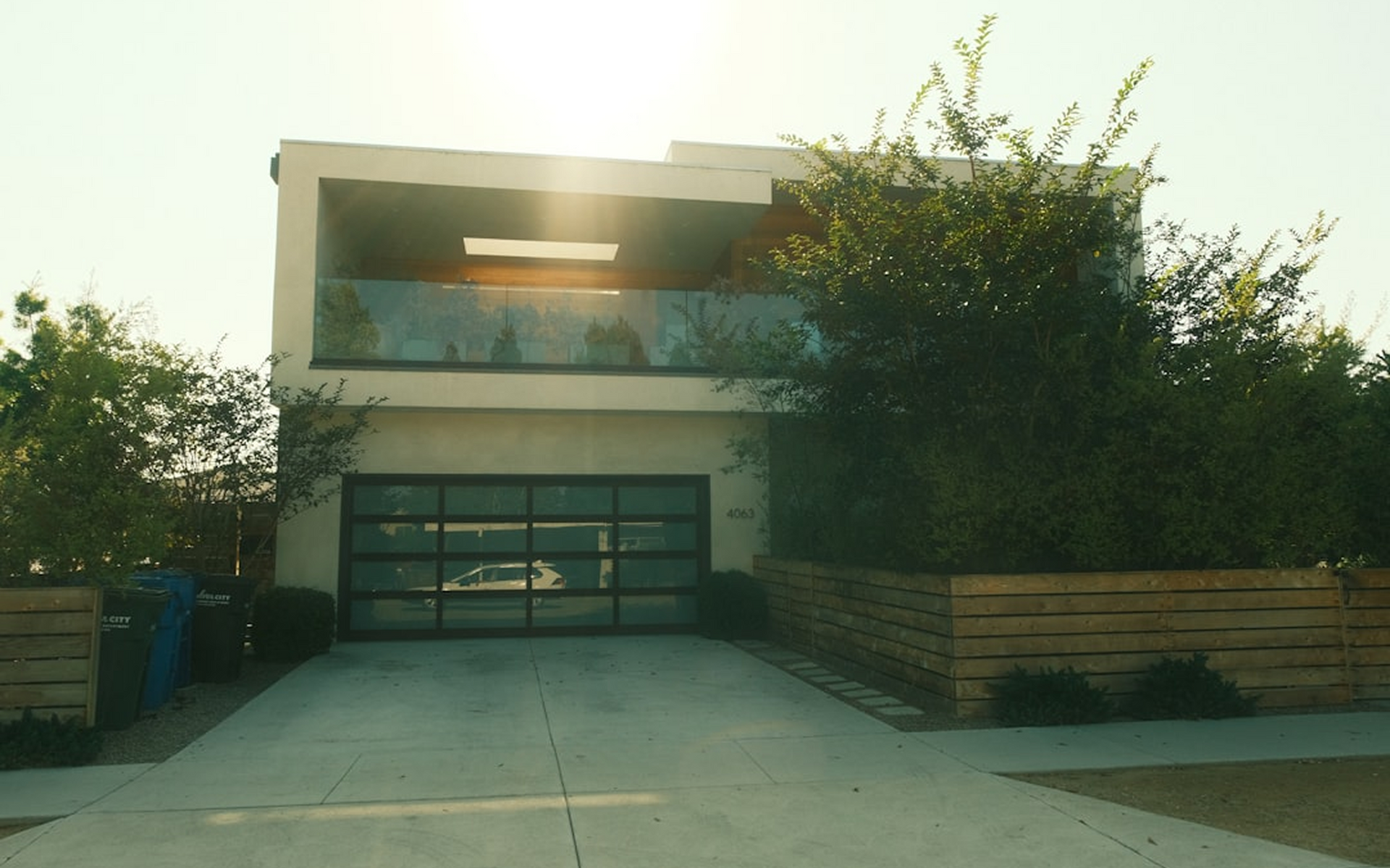 A modern, two-story house with a large glass garage door, a second-floor balcony, and a wooden fence at street level.