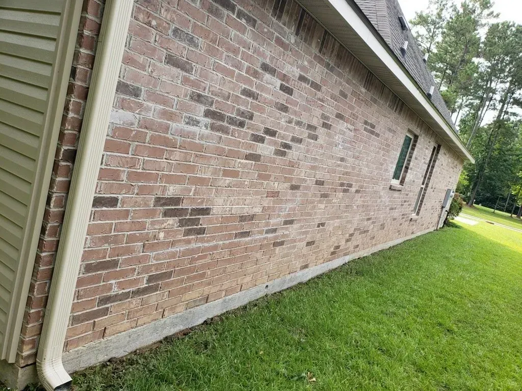 A side view of a brick exterior wall of a house with a beige downspout, concrete foundation, and green grass.