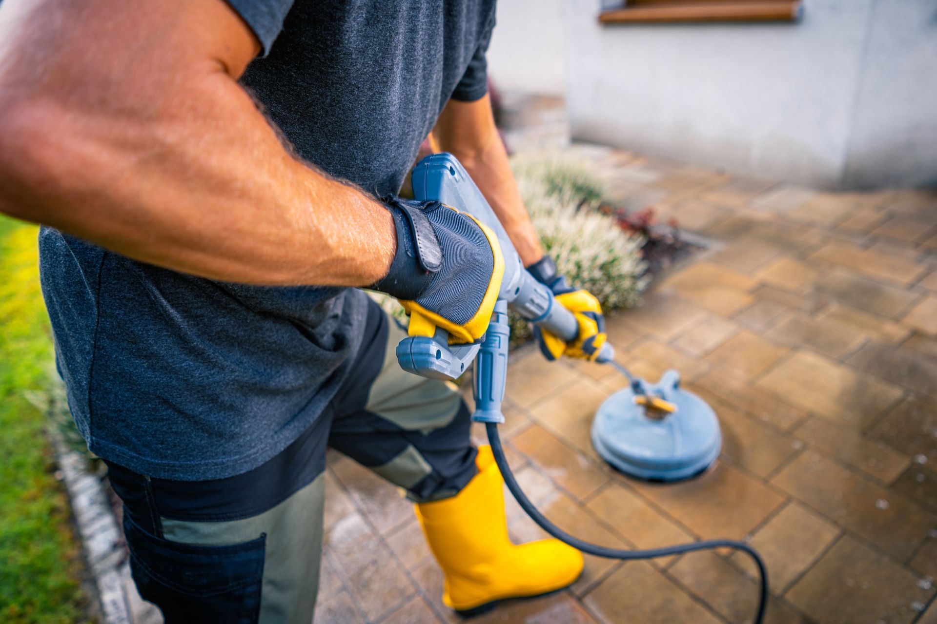 A person in work clothes and yellow boots uses a surface cleaner attached to a pressure washer on a paved patio.