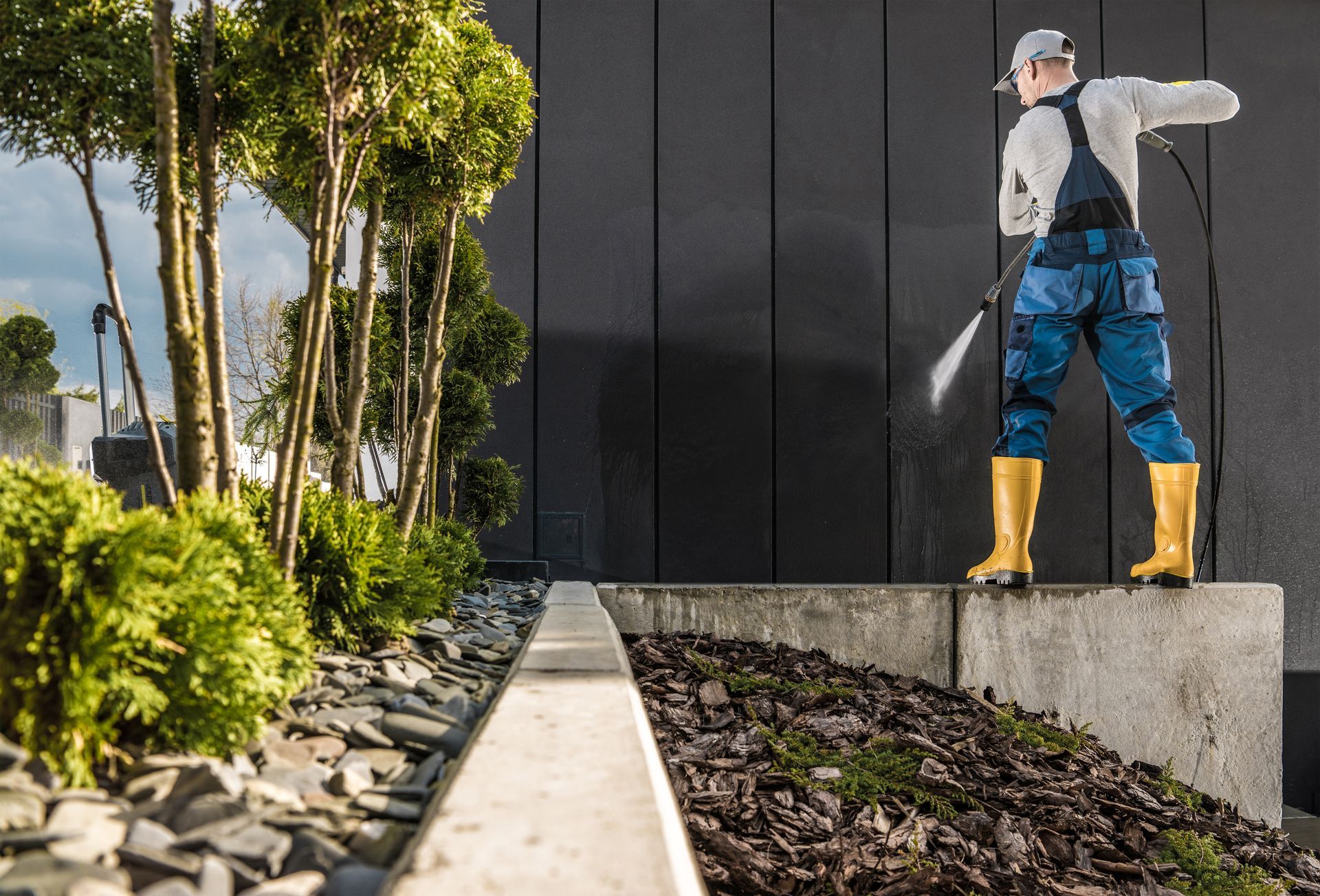 A person in protective gear and yellow boots uses a pressure washer to clean a dark wall outdoors near trees.
