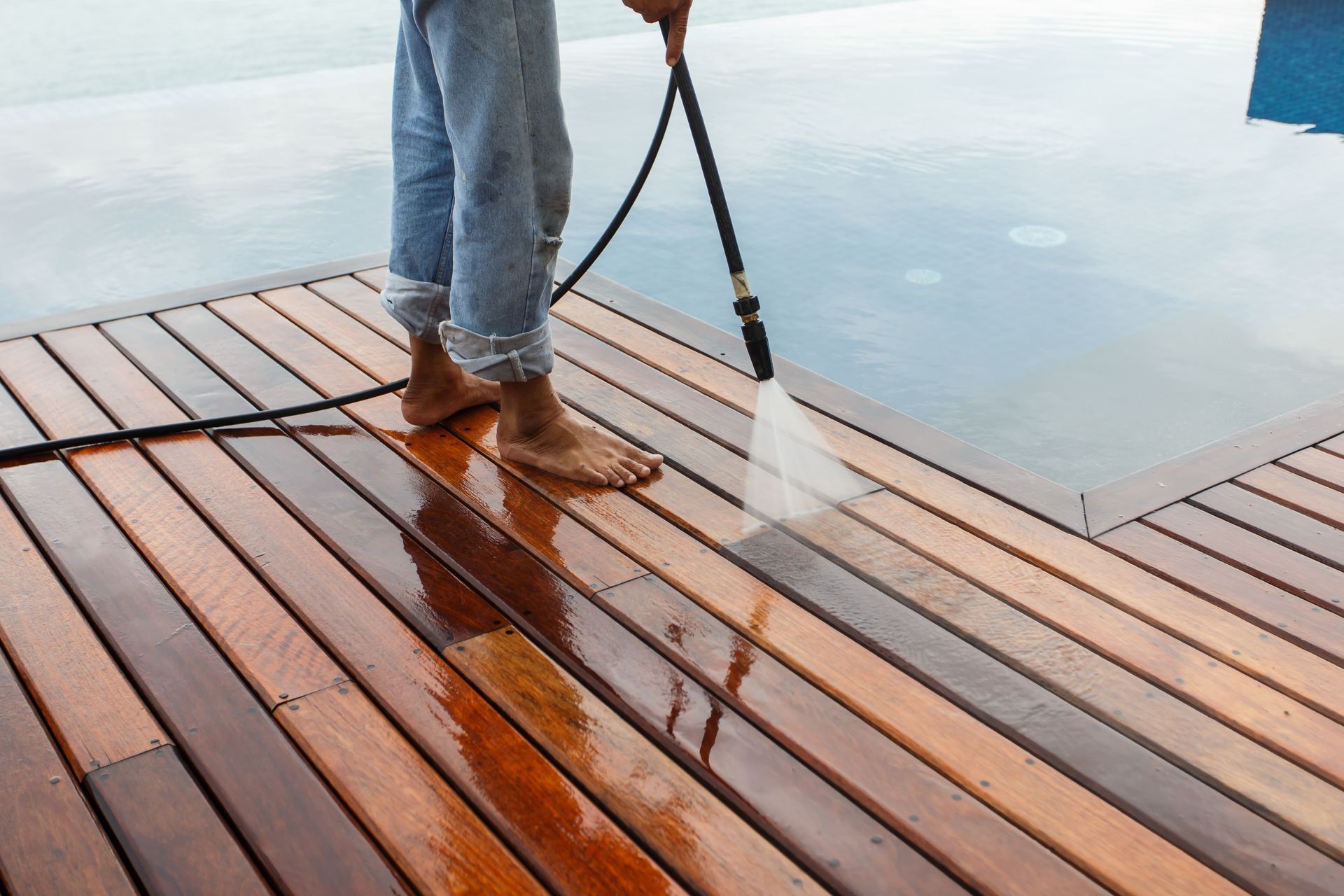 A person barefoot on a wooden deck by a pool, using a pressure washer to clean the planks.