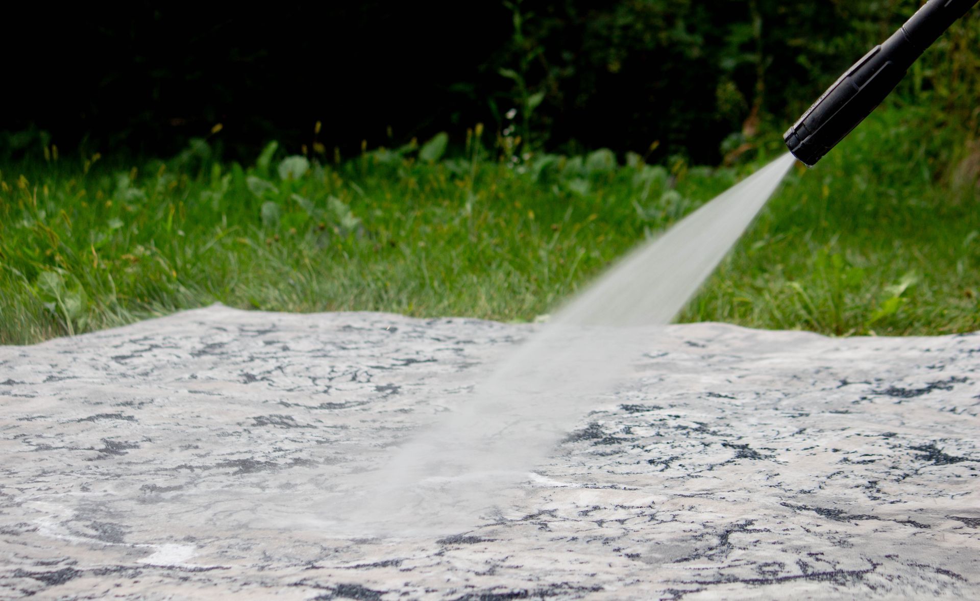 A high-pressure washer nozzle spraying a stream of water onto a stone surface outdoors near grass.