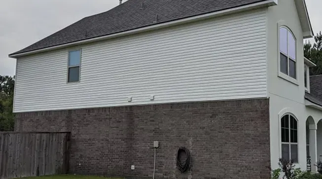 The side view of a two-story house with a white horizontal-sided upper level and a dark grey brick lower level.