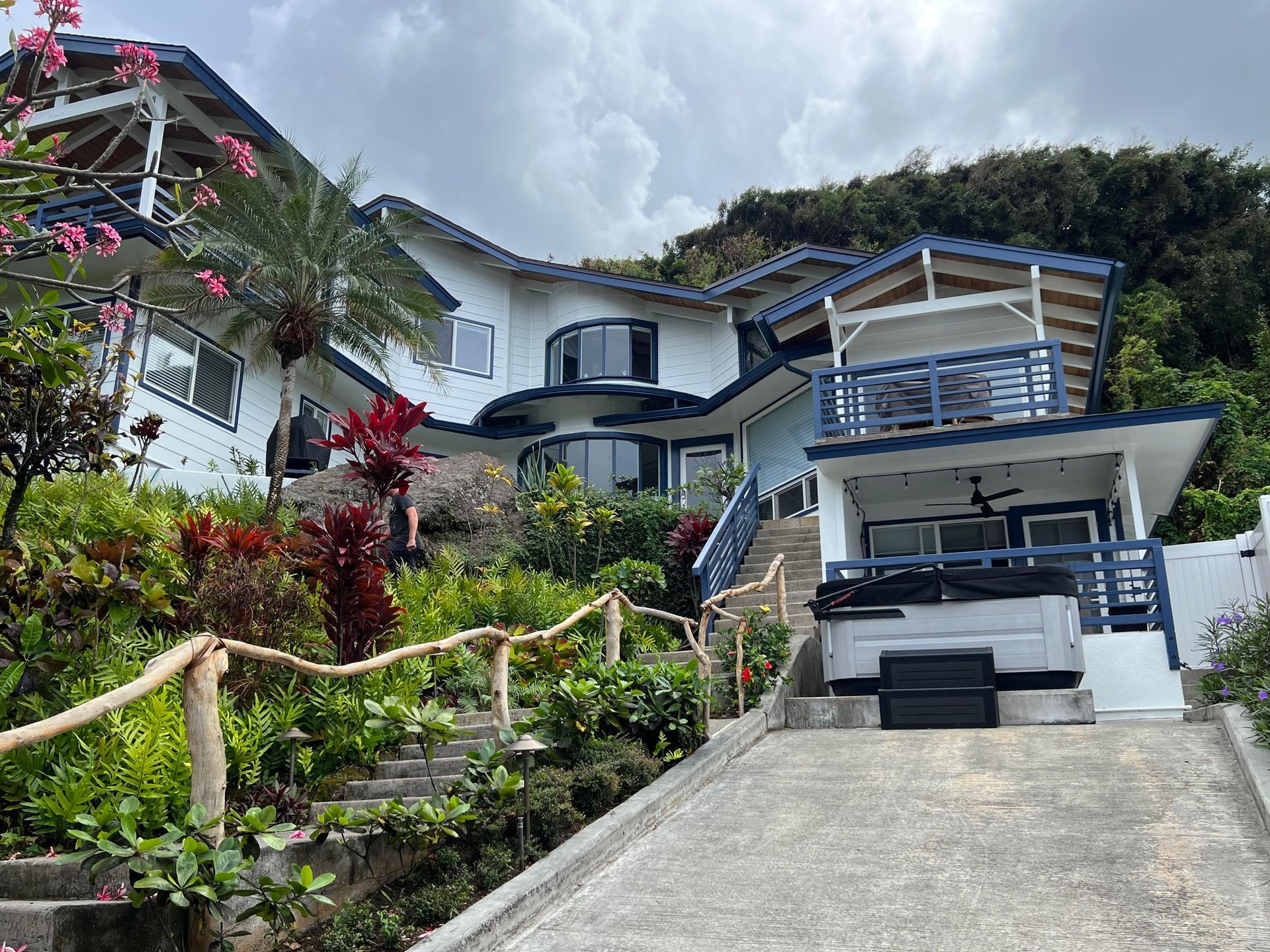 A white house with blue trim is nestled on a hillside amidst lush greenery, with a walkway leading to the entrance.