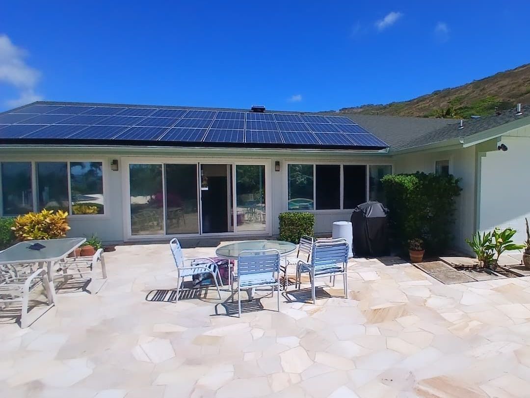 A house with solar panels on the roof, overlooking a patio with outdoor furniture under a bright blue sky.