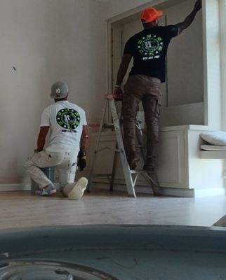 Two painters in work clothes are painting a room. One is on a ladder, the other is kneeling, both facing away.