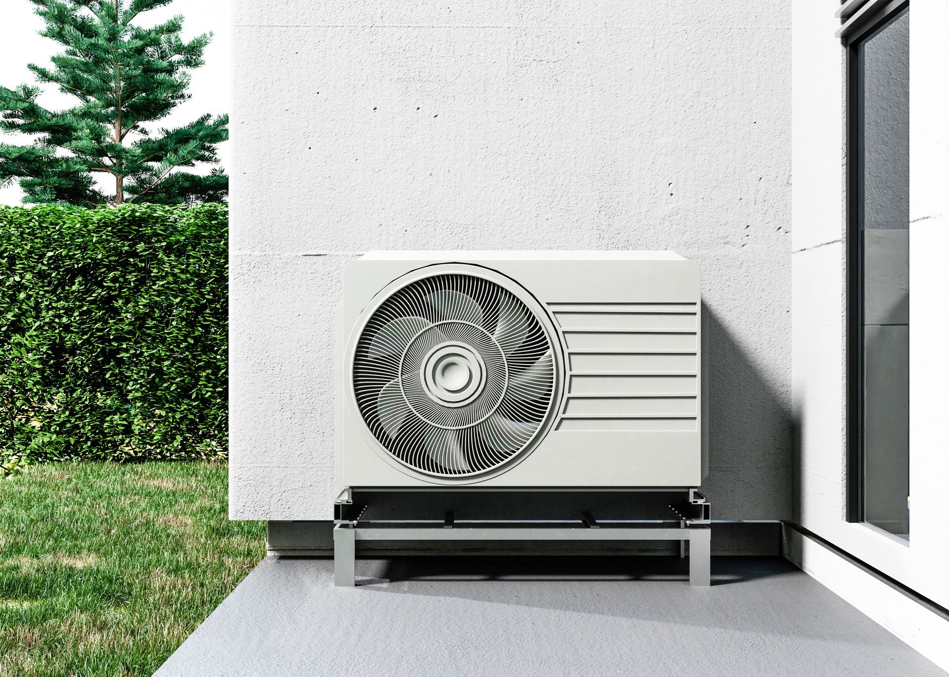White heat pump outside a building on a metal stand; green hedge and grass visible.