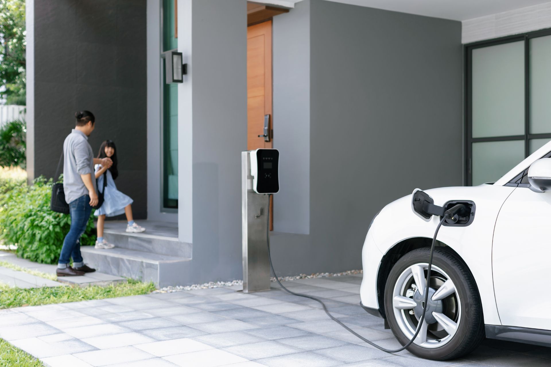 Father and daughter leaving a house, EV charging, gray exterior.