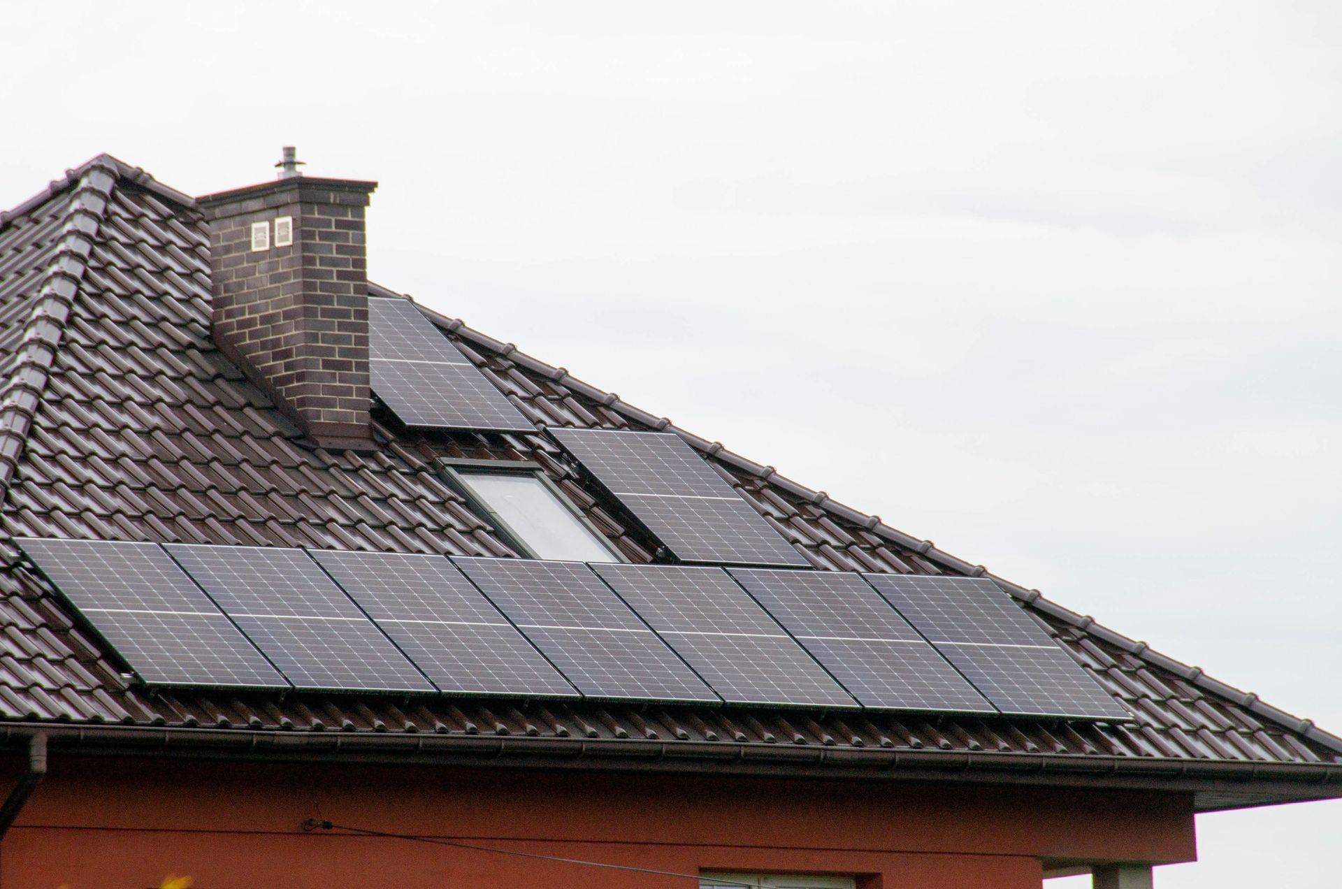 Solar panels on a tiled roof next to a brick chimney and a small window.