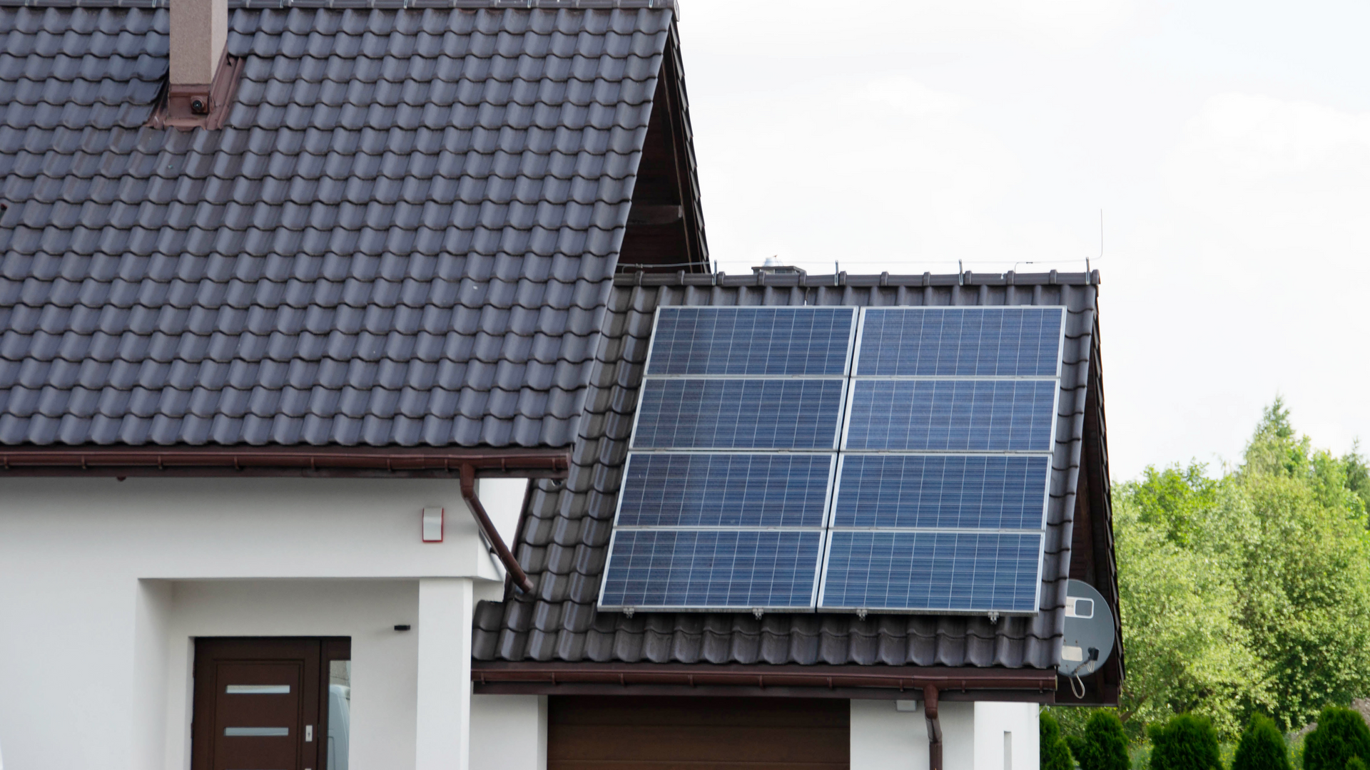 White house with solar panels on a dark roof. Brown door, green trees in background.