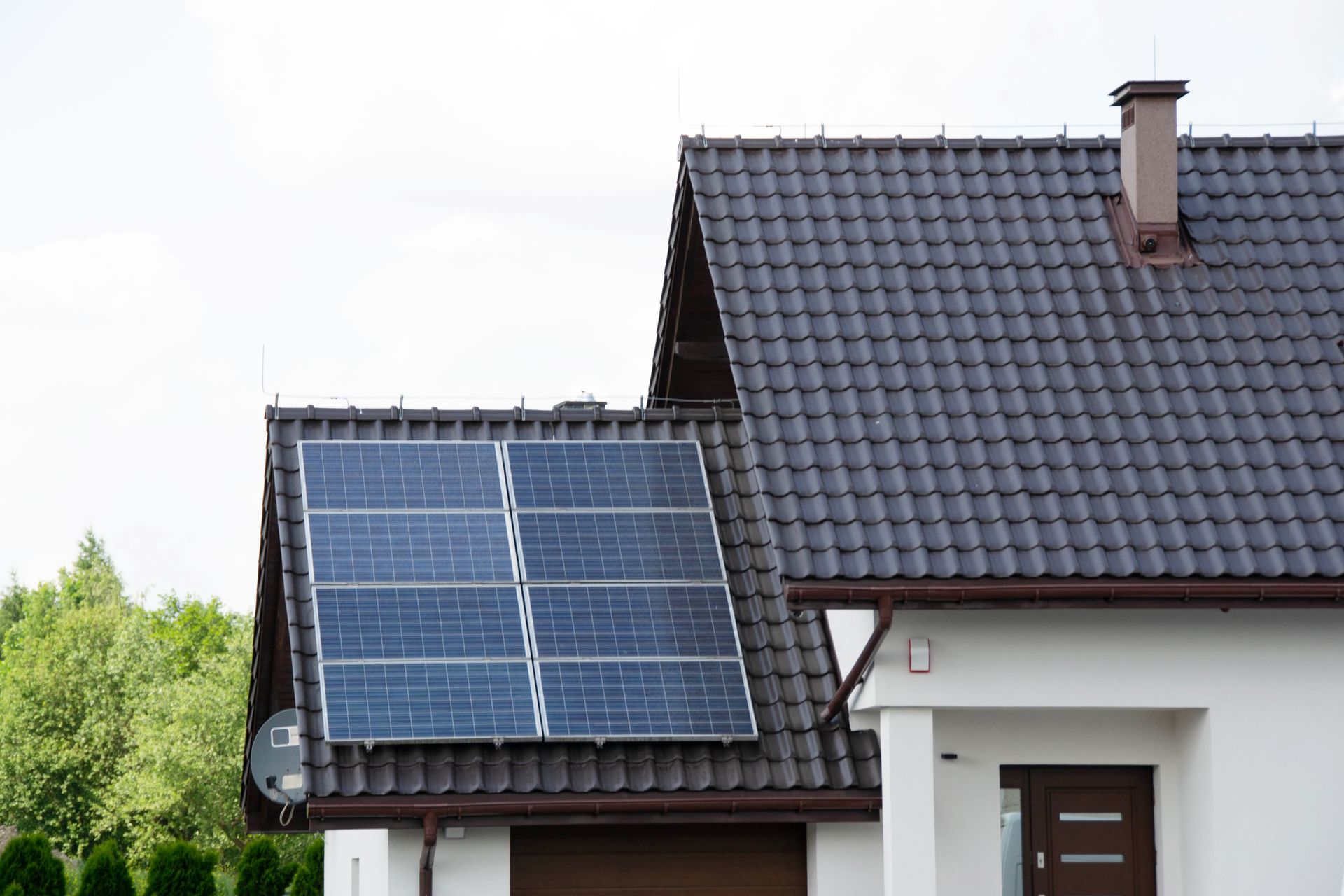 Solar panels on a house roof, white exterior, dark roof tiles, and green trees in the background.