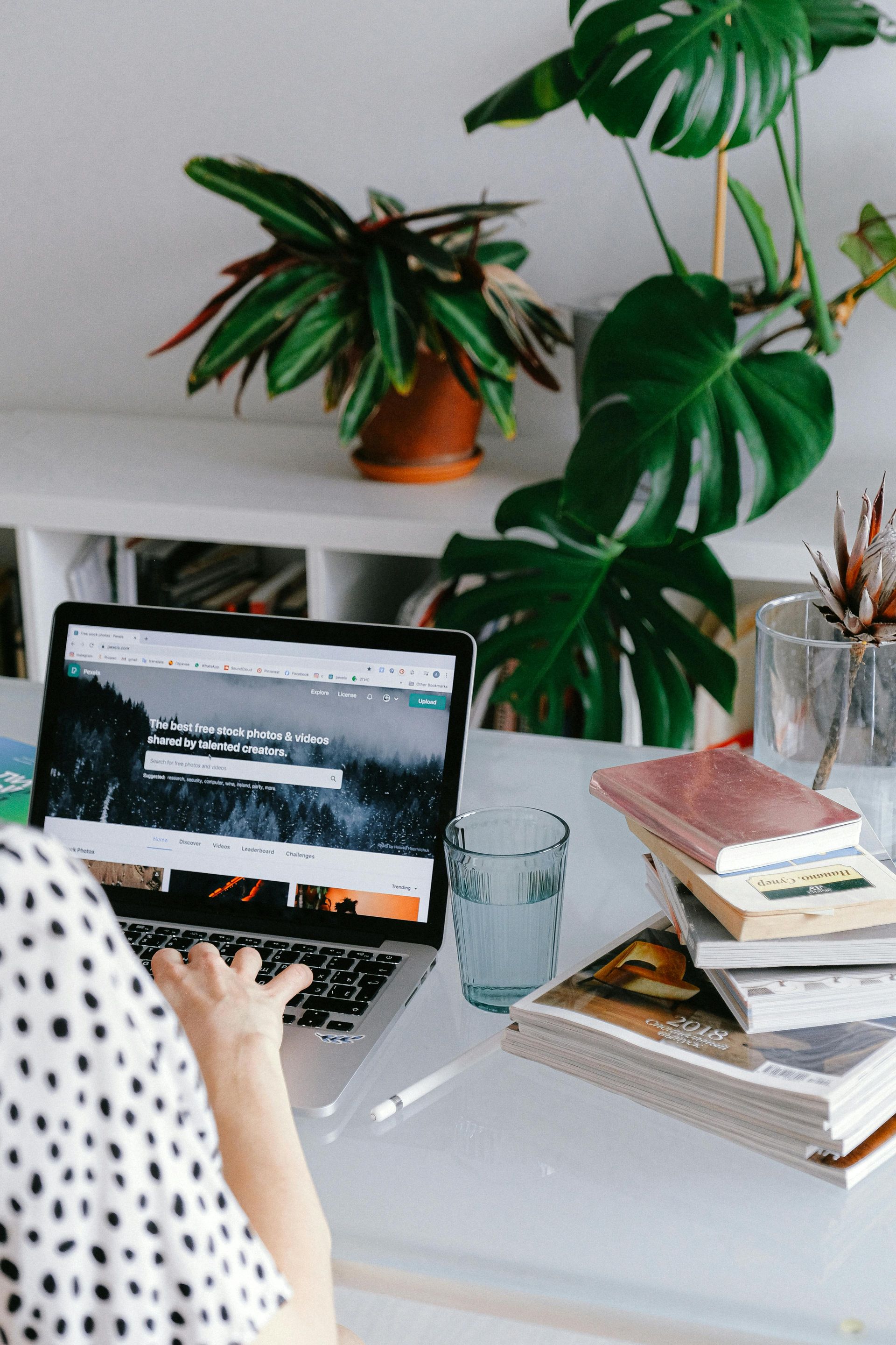 Personne tapant sur un ordinateur portable avec des plantes, des livres et un verre d'eau sur un bureau blanc.