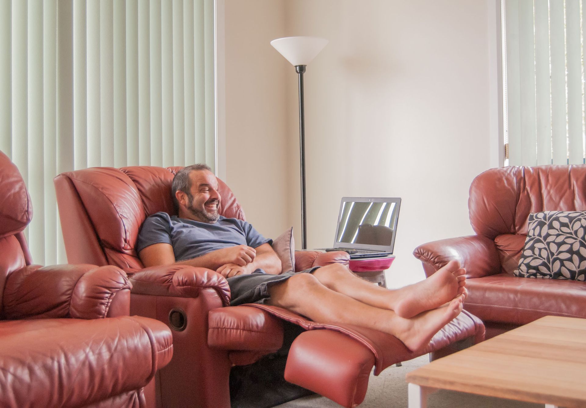 Man relaxing in a recliner with laptop, smiling. Living room with leather furniture, lamp, and window blinds. Man relaxing in a recliner with laptop, smiling. Living room with leather furniture, lamp, and window blinds.