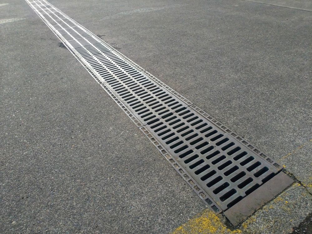 Long Metal Drainage Grate Embedded in Gray Asphalt Pavement — Hinch Plumbing In Breakwater, VIC