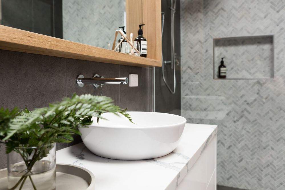 Modern bathroom with a white vessel sink, wood shelf, mirror, and gray tiled wall — Hinch Plumbing In Breakwater, VIC
