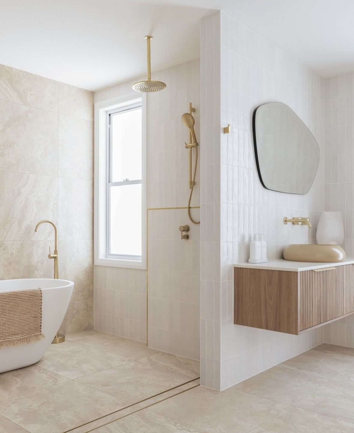 Minimal beige bathroom with freestanding tub, window shower, and floating wood vanity with oval mirror — Hinch Plumbing In Breakwater, VIC