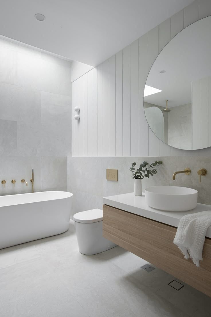 Minimalist white bathroom with freestanding tub, wood vanity, round mirror, and brass fixtures.