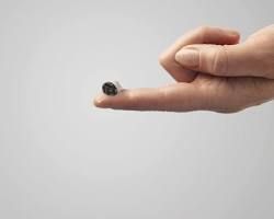 Mano che tiene un piccolo apparecchio acustico scuro sulla punta del dito, su uno sfondo neutro.